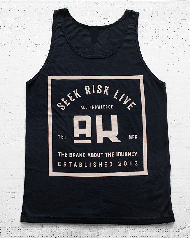 A black sleeveless tank top laid flat against a white textured brick wall. The front has a large beige rectangular graphic with the words 'SEEK RISK LIVE', a large 'AH' in the center, and smaller text below reading 'ALL KNOWLEDGE', 'THE BRAND ABOUT THE JOURNEY', and 'ESTABLISHED 2013'.