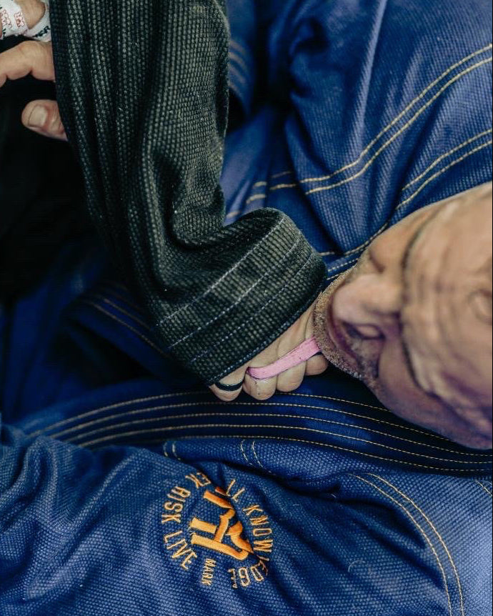 Two individuals in a grappling position on a blue mat with one wearing a blue gi.